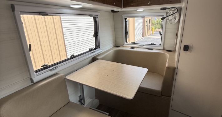 Interior of a small RV dining area with beige cushioned bench seats around a light wood table, dark wood overhead cabinets, and two windows with blinds letting in daylight.
