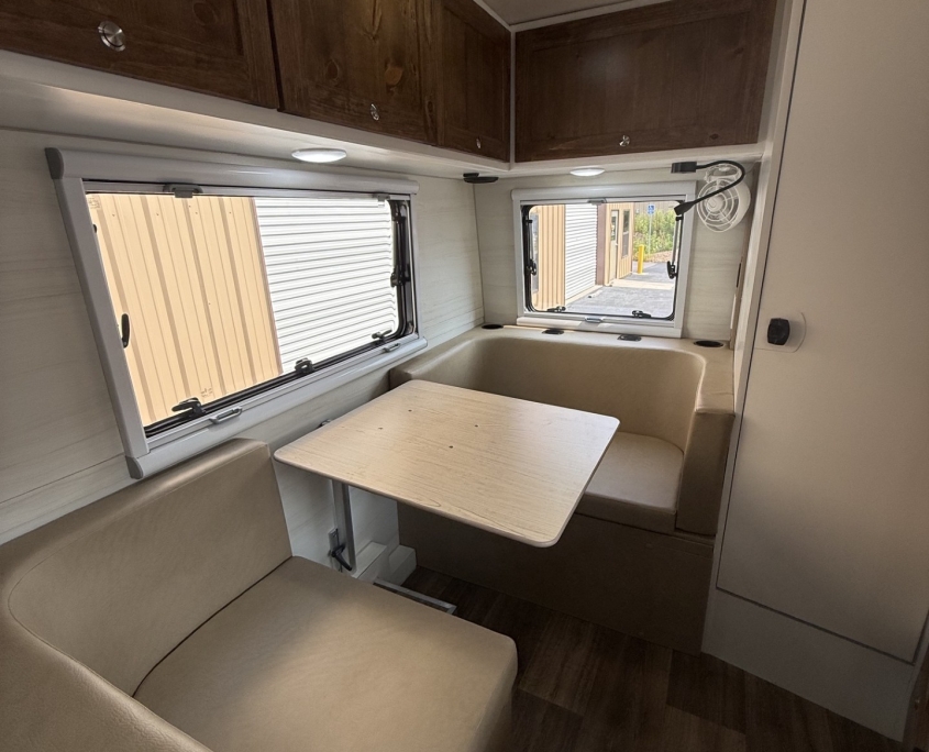 4e3e9057-6949-42d7-aa3e-61298c91cfd1 | Tiger Adventure Vehicles RV dinette area with beige bench seating, light wood table, and dark wood overhead cabinets; window with blinds behind.