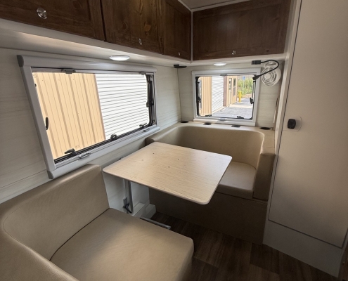 Interior of a camper or RV dining area: beige booth seating around a light wood table, dark wood overhead cabinets, and two windows with blinds letting in daylight.