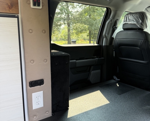 Camper van interior with a beige padded pillar, control panel, and a side window showing trees outside.