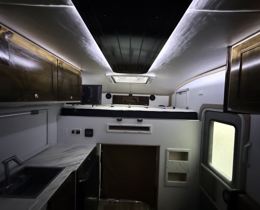 Interior of a compact RV kitchen with marble counters, wooden cabinets, and a sink on the left; ceiling vent above.