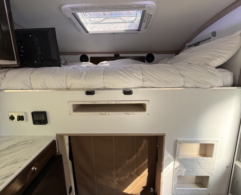 Bunk bed in a small travel trailer with a white comforter, a ceiling hatch window above, and built-in storage nearby.
