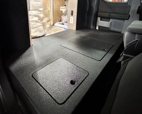 Flat, black textured vehicle cargo floor with two rectangular access panels; one panel shows a small circular cutout.