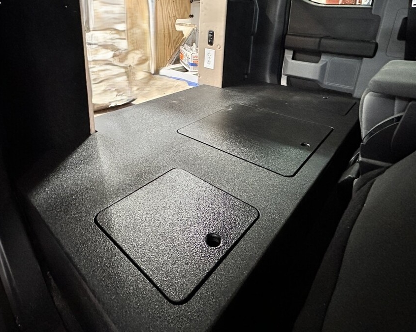 Hatches on Extended Cab Truck | Tiger Adventure Vehicles Flat, black textured vehicle cargo floor with two rectangular access panels; one panel shows a small circular cutout.