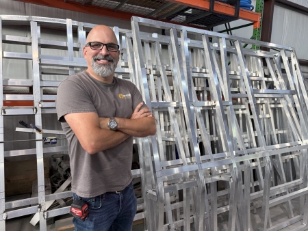 James B | Tiger Adventure Vehicles Smiling man with glasses and a beard stands with arms crossed among aluminum framing in a workshop, posing for the photo.