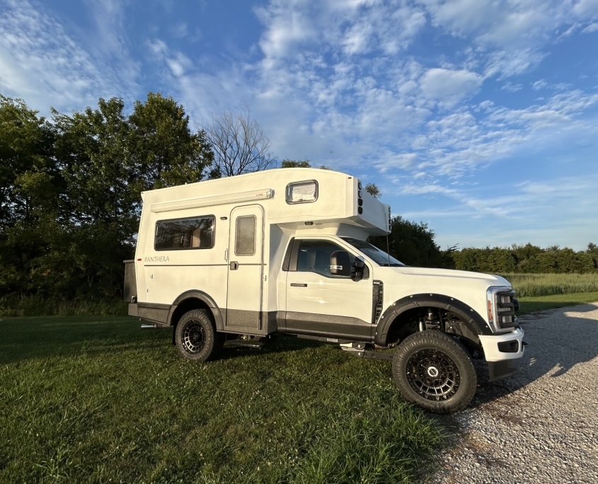 White Panthera motorhome with a high roof and large windows parked on grass beside a gravel road, under a blue sky with scattered clouds.