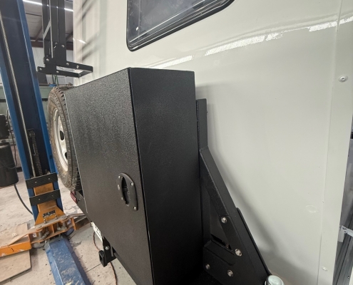 Industrial workshop scene: a large vehicle on a hydraulic lift, side window visible, with a textured black mounting platform attached to the chassis