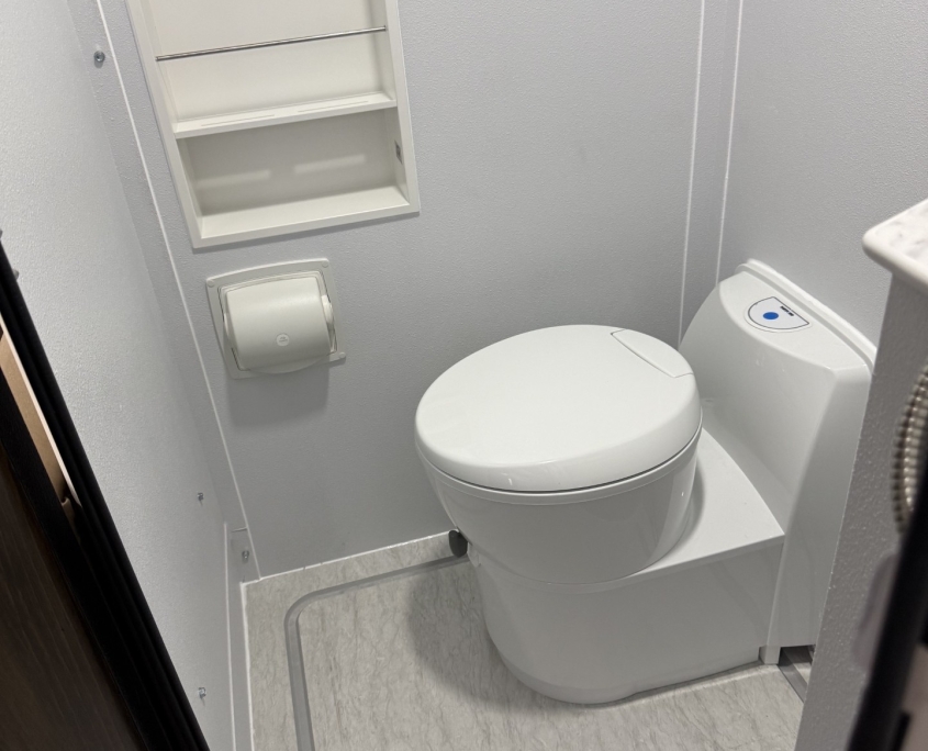 Well draining shower pan in the Tiger RVs | Tiger Adventure Vehicles Small white bathroom with a modern toilet, wall cabinet with shelves, and a wall-mounted toilet paper dispenser on light walls, compact design.