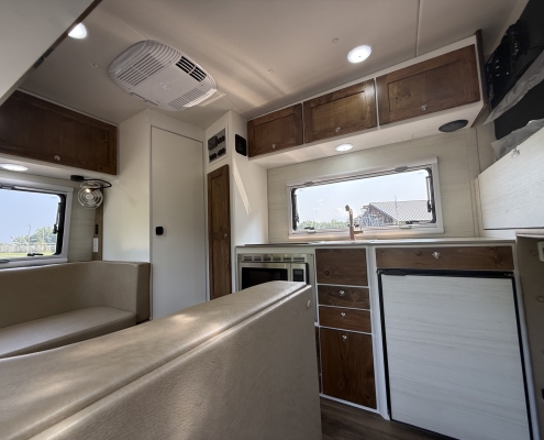 Interior of a camper van/kitchen area with beige seating, white cabinets, and brown wood upper cabinets along a wall stove and appliances nearby