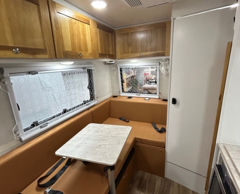 Cozy camper van interior with brown cushioned bench seating, a small table, and wooden overhead cabinets above large windows.