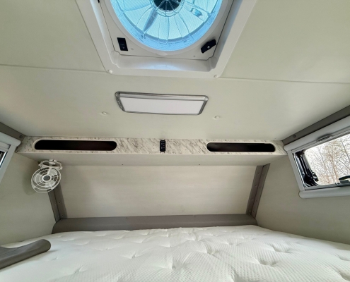 Interior of a camper van sleeping area with a round roof vent and overhead light panel above a padded bed