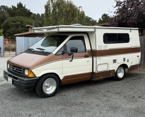 b7473adf-a930-48ee-94f5-0483021b569d | Tiger Adventure Vehicles | Tiger Adventure Vehicles Brown and cream camper van with pop-up roof extended, parked on a gravel lot nearby a wooden fence.