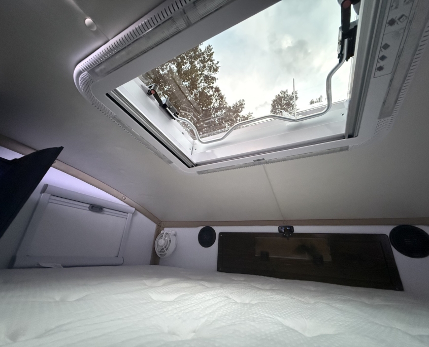 Open skylight above a bed in an RV, with a curved metal handle and trees visible outside