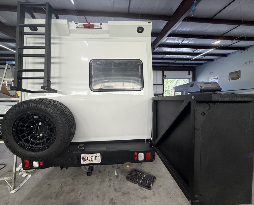 Rear of a white utility trailer on a black trailer, with a spare tire mounted on the back and a tall ladder on the left, inside a workshop in progress.