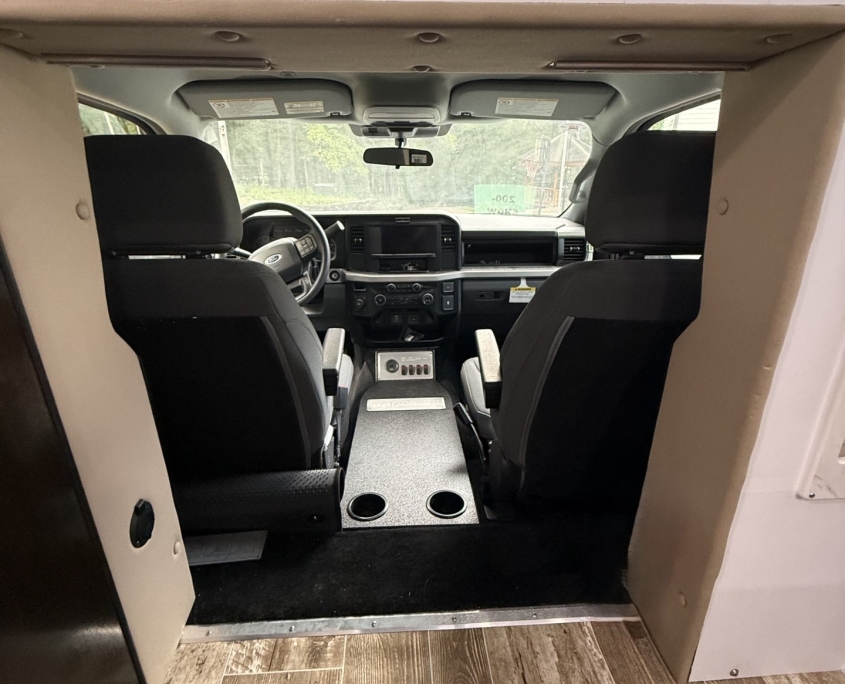 Interior of a vehicle cabin viewed from the back, showing two front seats, a central console, and a wide dashboard. Wooden-look flooring in the foreground.