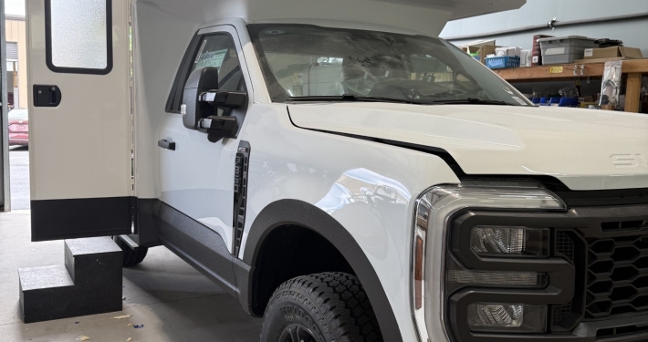 White utility van with a large beige bucket lift mounted over the cab, door open, inside a workshop.