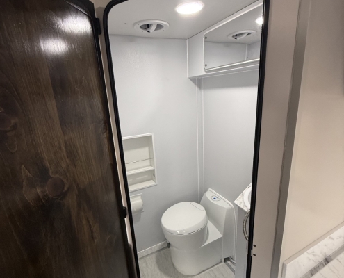 Small bathroom in a compact space showing a white toilet, sink, and wall shelf with an oval mirror; gray walls and ceiling vent visible in a compact, RV-like bathroom.
