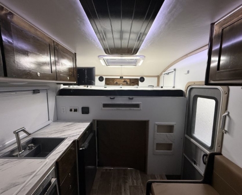 Interior of a camper van/kitchenette: marble counter, sink, dark wood cabinets, and compact appliances with a seating area by a door with a window on the right.