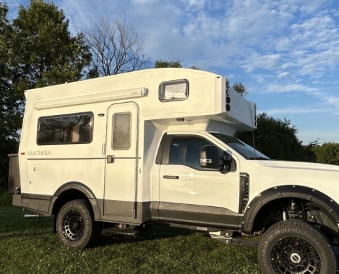White Panthera camper van with raised roof, parked on a grassy field under a blue sky with trees nearby.