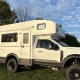 White Panthera camper van with raised roof, parked on a grassy field under a blue sky with trees nearby.