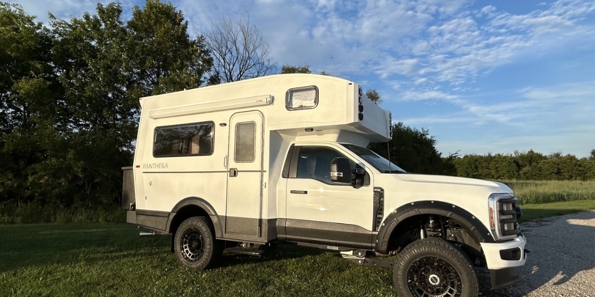 White Panthera camper van with raised roof, parked on a grassy field under a blue sky with trees nearby.