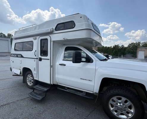39a20657-345b-4ac1-b5da-64644542dbae | Tiger Adventure Vehicles | Tiger Adventure Vehicles White pickup truck with a raised camper over the cab, labeled 'BENGAL 4X4' on the side, parked in a lot under a blue sky.