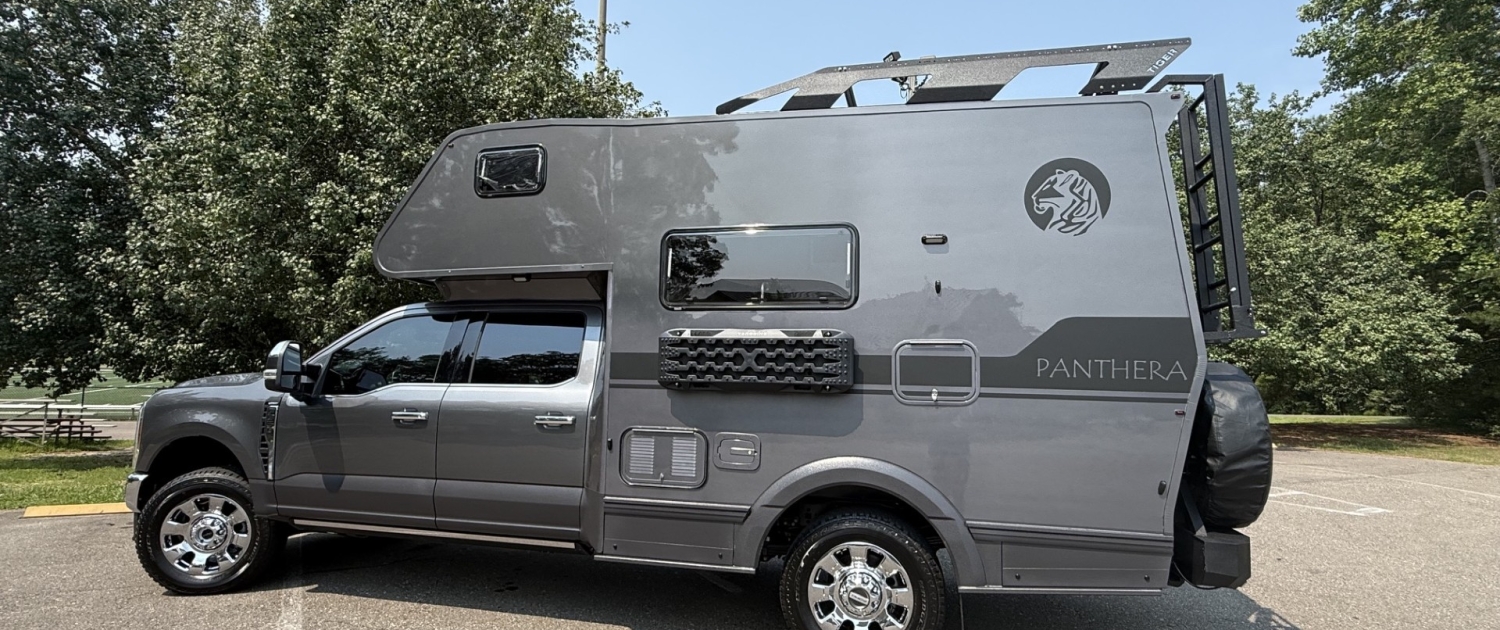 | Tiger Adventure Vehicles Grey Panthera motorhome parked in a lot, showing a rooftop rack and lion logo on the side behind the cab.