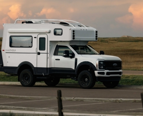 White Overland camper truck (Ford) parked on a road with a spacious desert-like plain, tail in the distance, sunset sky in the background.