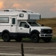 Panthera200-compact-main_img | Tiger Adventure Vehicles White Overland camper truck (Ford) parked on a road with a spacious desert-like plain, tail in the distance, sunset sky in the background.