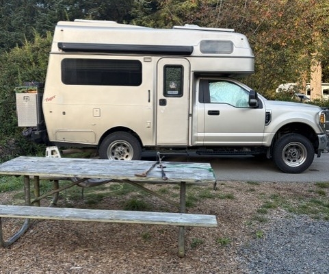 6864690f-7302-4be2-bb01-b084c21f75eb | Tiger Adventure Vehicles | Tiger Adventure Vehicles Beige camper van with a raised roof parked on a gravel lot, viewed from the side; a weathered wooden picnic table and a green camping chair sit in the foreground, with trees in the background.