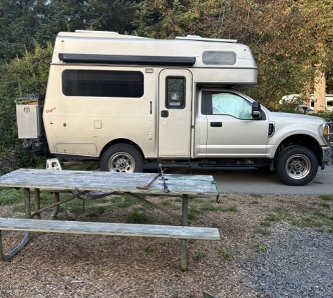 6864690f-7302-4be2-bb01-b084c21f75eb | Tiger Adventure Vehicles Beige camper van with a raised roof parked on a gravel lot, viewed from the side; a weathered wooden picnic table and a green camping chair sit in the foreground, with trees in the background.