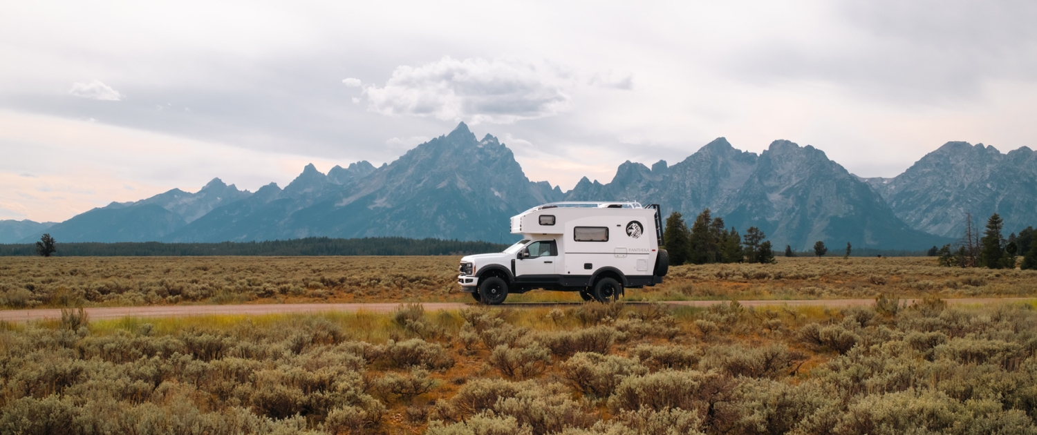 Kate-and-Joe-TIGER-5289 | Tiger Adventure Vehicles White camper van traveling through a desert plain with rugged mountains in the distance under a cloudy sky