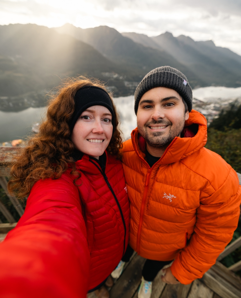 Katie and Joe-Juneau | Tiger Adventure Vehicles Smiling couple takes a selfie in red and orange jackets on a wooden overlook, with mountains and a lake in the background.