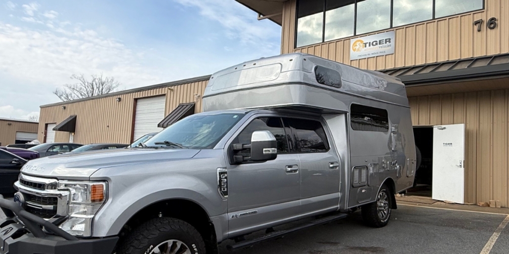 Panthera200-compact-main_img | Tiger Adventure Vehicles | Tiger Adventure Vehicles Silver pickup truck with a raised overcab camper parked outside a tan industrial building with a Tiger Vehicles sign above the door.