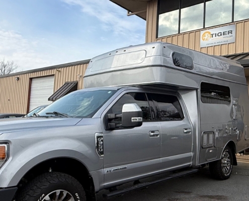 Silver pickup truck with a raised overcab camper parked outside a tan industrial building with a Tiger Vehicles sign above the door.