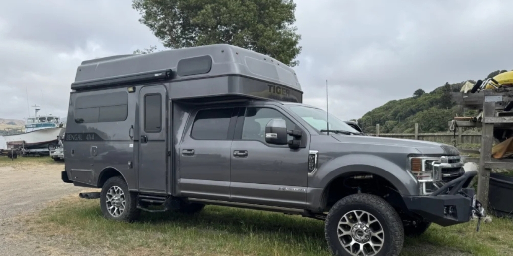 Panthera200-compact-main_img | Tiger Adventure Vehicles | Tiger Adventure Vehicles Grey pickup with a high-roof camper shell parked on a grassy area, with boats and a fence nearby on a cloudy day.