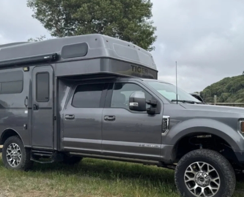 Grey pickup with a high-roof camper shell parked on a grassy area, with boats and a fence nearby on a cloudy day.