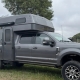tiger-marketplace-consignment-main_img | Tiger Adventure Vehicles Grey pickup with a high-roof camper shell parked on a grassy area, with boats and a fence nearby on a cloudy day.