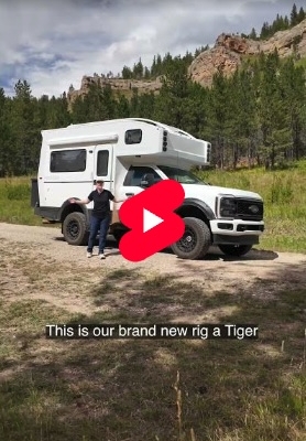 Video thumbnail: white camper truck with raised living quarters; two people stand beside it in a grassy field with mountains in the background, play button overlay.