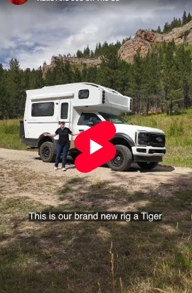 yt-featured-img-placeholder-2 | Tiger Adventure Vehicles Video thumbnail: white camper truck with raised living quarters; two people stand beside it in a grassy field with mountains in the background, play button overlay.