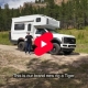 Video thumbnail: white camper truck with raised living quarters; two people stand beside it in a grassy field with mountains in the background, play button overlay.